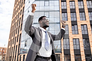 Excited african american businessman celebrating success and raising hands up, standing outdoors near office center