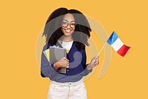 Excited african american lady student with copybooks, holding flag of France, posing on yellow background