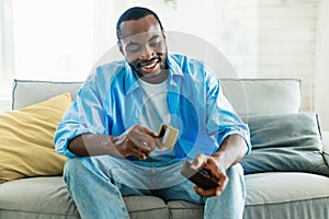 Excited african american man selecting credit card from his card wallet, sitting on sofa in living room interior