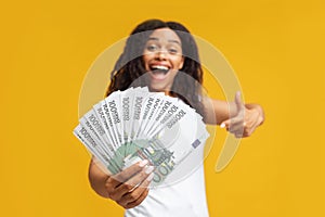 Excited african american woman holding european money cash pointing finger at banknotes, standing over yellow background