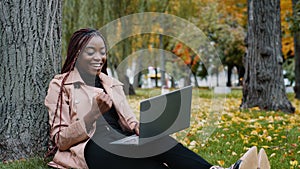 Excited african american woman sitting in autumn park playing online game on laptop gambling competition happy young