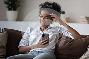 Excited African American woman using smartphone, sitting on cozy couch