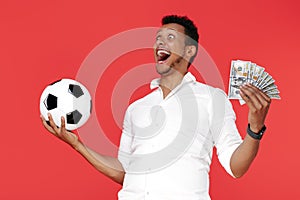 Excited african american young man holding cash money and soccer ball over red background.