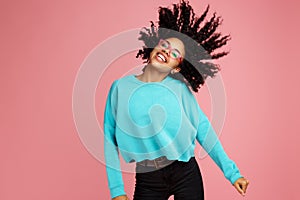 Excited african american young woman with bright smile dressed in casual clothes, glasses and headphones dance over pink