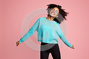 Excited african american young woman with bright smile dressed in casual clothes, glasses and headphones dance over pink