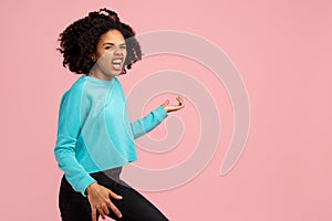 Excited african american young woman with bright smile dressed in casual clothes, glasses and headphones dance over pink