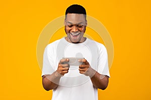 Excited African Guy Using Smartphone Playing Mobile Games, Yellow Background