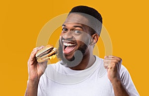 Excited African Man Eating Burger Gesturing Yes Over Yellow Background