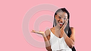 Excited Afro woman in stylish dress touching her face, showing something on empty space over pink background