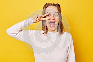 Excited amazed teenage girl in jumper with ponytails isolated over yellow background celebrating her victory success showing v