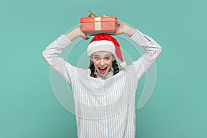 Excited amazed teenager girl in Santa Claus hat holding present box above her head, keeps mouth open