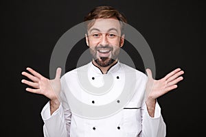 Excited amazed young bearded male chef cook or baker man in white uniform shirt posing isolated on black wall background