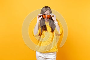 Excited amazed young woman in fur sweater, white pants holding and taking off heart orange glasses isolated on bright