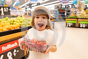 Excited american kid with shopping trolley with in grocery store. Little boy in the supermarket holdind strawberry.