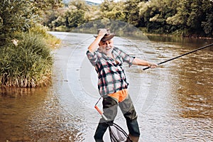 Excited angler senior man fisherman in cowboy hat with fishing rod, spinning reel on river. Old man catching fish