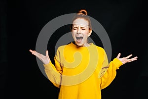 Excited angry young woman in yellow sweater screaming with closed eyes on isolated black background