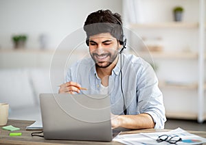 Excited arab man in headphones studying online with laptop at home, watching webinar and taking notes to notepad