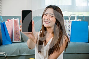 Excited Asian buying woman smiling showing mobile phone with many shopping bags at home