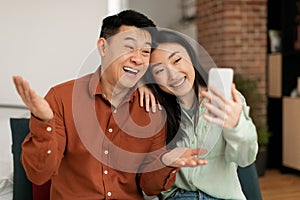 Excited asian couple taking selfie on smartphone, communicating via video call, talking and smiling at webcamera