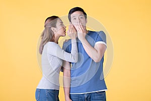 Excited Asian couple whispering secrets to each other isolated on yellow background
