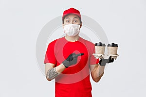 Excited asian delivery man in red cap, medical mask and gloves, safely deliver your beverege, holding cups of beverage