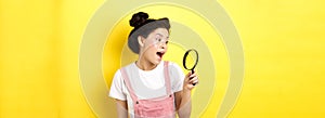 Excited asian girl found interesting thing, looking through magnifying glass amazed, standing on yellow summer