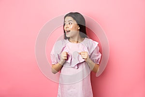 Excited asian girl look left with motivated face, smiling happy, standing in dress on pink background