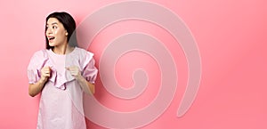 Excited asian girl look left with motivated face, smiling happy, standing in dress on pink background