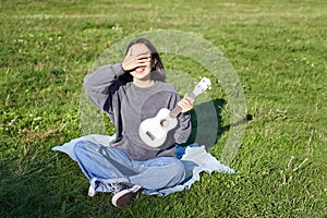 Excited asian girl sits in park with ukulele, plays instrument and feels happy upbeat, positive moments and people