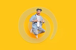 Excited asian man in stylish outfit showing thumb ups while jumping on yellow studio background, recommend something