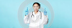 Excited asian medical worker, female doctor pointing fingers up, wearing sterile gloves and uniform, standing over blue