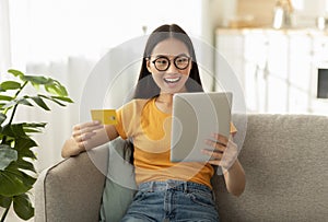 Excited asian woman shopping from home, using digital tablet and credit card, buying things in web store