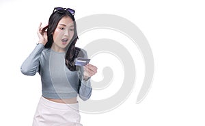 Excited asian woman with sunglasses looking credit card in hand standing on white background. Cheerful surprised young girl