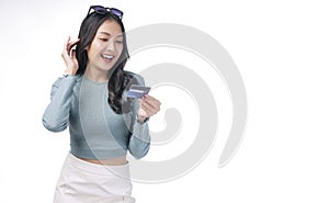 Excited asian woman with sunglasses looking credit card in hand standing on white background. Cheerful surprised young girl