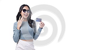 Excited asian woman wear sunglasses looking credit card in hand standing on white background. Cheerful surprised young girl