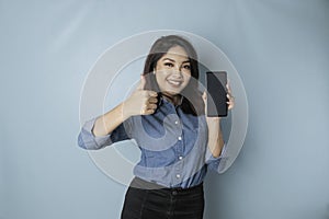 Excited Asian woman wearing a blue shirt gives thumbs up hand gesture of approval while holding her smartphone, isolated by blue