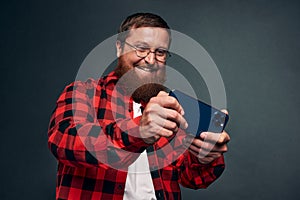 Excited bearded man in plaid shirt playing on smartphone over grey background