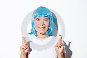 Excited beautiful asian girl in blue wig biting lip and looking hopeful at camera, making wish with fingers crossed