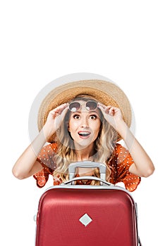 excited beautiful girl holding sunglasses and sitting near travel bag