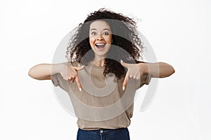 Excited beautiful woman showing announcement, pointing fingers down at advertisement and smiling amazed, happy to