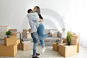 Excited black couple celebrating moving day, guy lifting his wife in their new apartment, standing among boxes