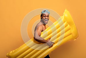 Excited black guy in snorkeling mask holding inflatable lilo, walking to beach on yellow studio background