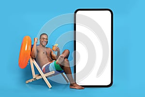 Excited black guy with tropical cocktail chilling in lounge chair near huge smartphone with blank screen for your ad