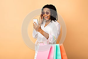 Excited Black Lady Using Phone Holding Shopper Bags, Beige Background photo