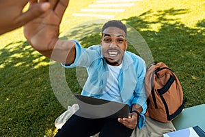 Excited black male student giving high five to friend while using laptop computer, sitting on grass in park outside