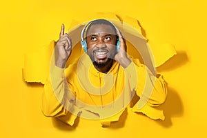 Excited black man enjoying music via wireless headphones and dancing in paper hole against yellow background