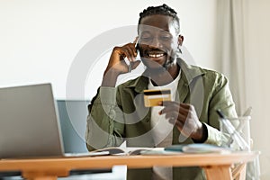 Excited black man holding credit card, having phone conversation with bank representative, sitting at home