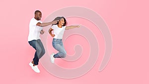 Excited black man and woman jumping up in the air and pointing at free space, pink studio background, panorama