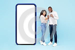 Excited black spouses standing near big smartphone with empty screen, using gadget and shaking clenched fists, mockup