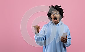 Excited black teen guy using cellphone, advertising cool application, pointing aside at blank space on pink background